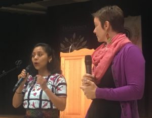 A Guatemalan woman in traditional Mayan dress, holding a microphone and gesturing, while a white, female colleage, her translator, looks on.