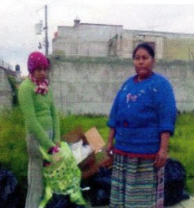 a teenaged girl holding a plastic bag, in front of a small pile of what appears to be garbage, including a cardboard box. On the right side of the photo is an older Maya woman in traditional dress. 
